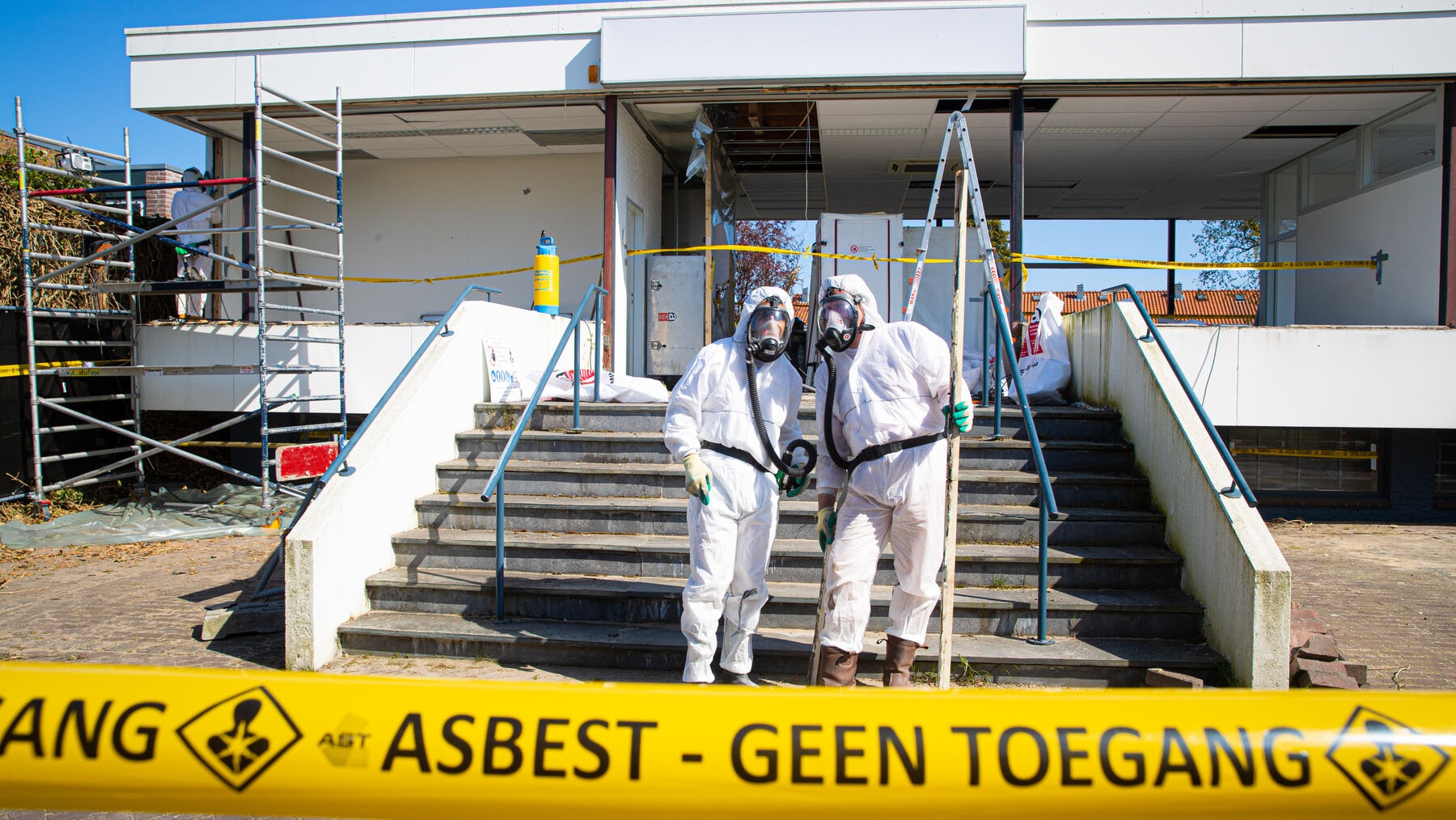 Saneerders in witte beschermende overall en masker verwijderen asbesthoudende golfplaten van een dak