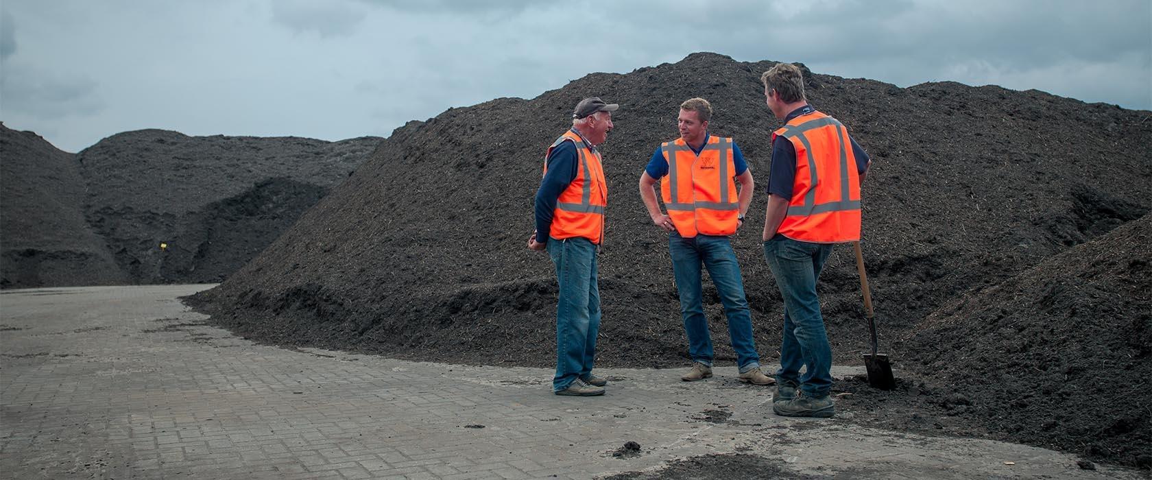 Drie medewerkers van Van Werven in oranje veiligheidsvest in gesprek voor lange bergen compost op een recyclinglocatie