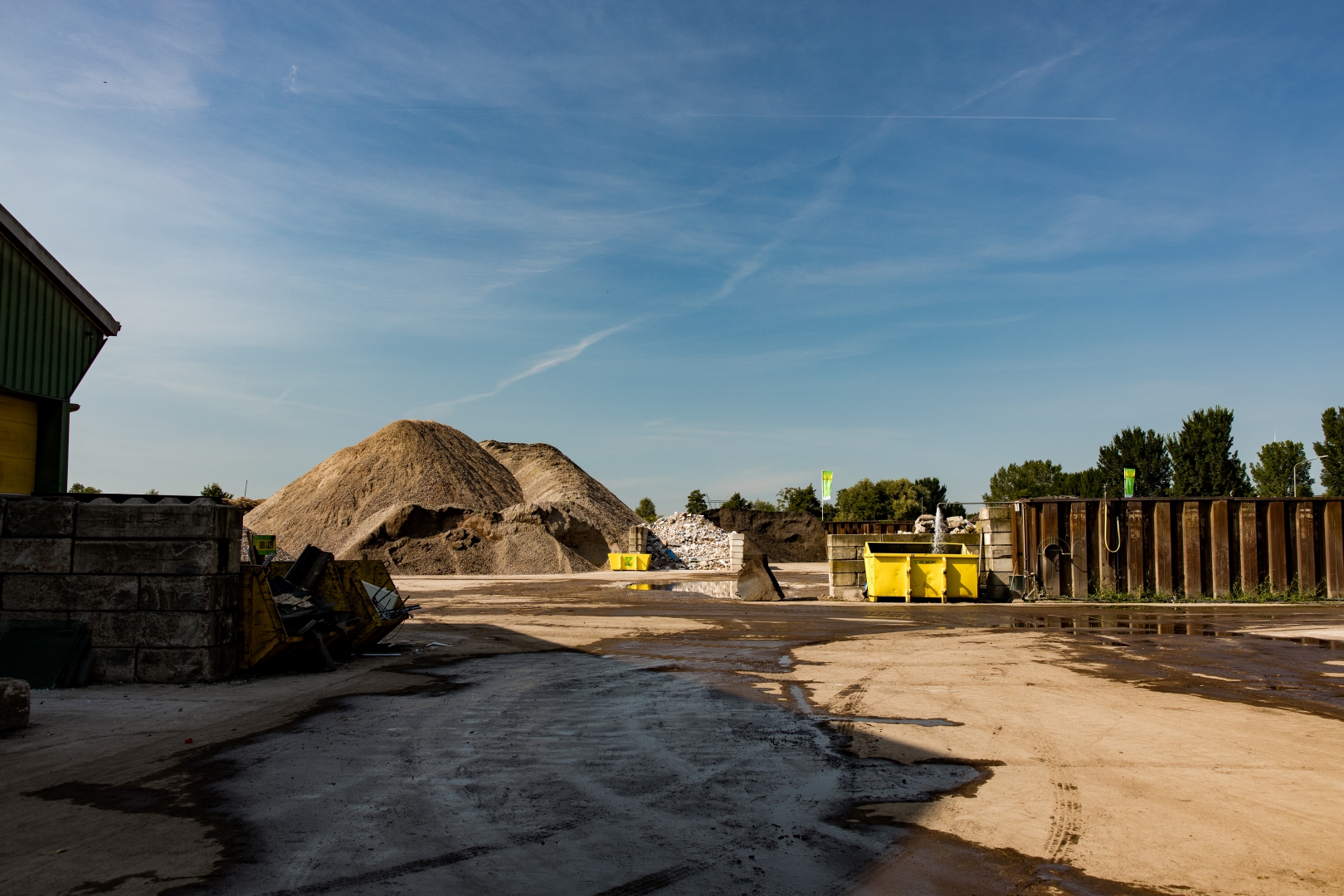 Het terrein van de recyclingvestiging onder een blauwe lucht, met een berg zand, gele containers en een damwand langs de rand