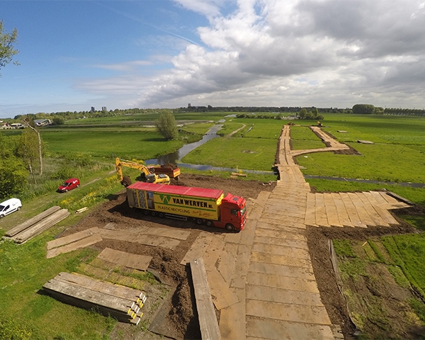 Luchtfoto van een tijdelijke rijbaan van houten rijplaten door een polder, met een vrachtwagen van Van Werven erop en een kraan die platen legt