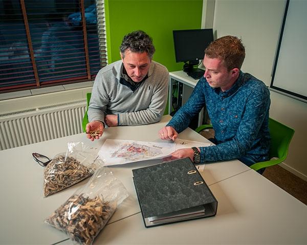Twee medewerkers bekijken aan een tafel een tekening, met zakken houtchips en boomschors ernaast