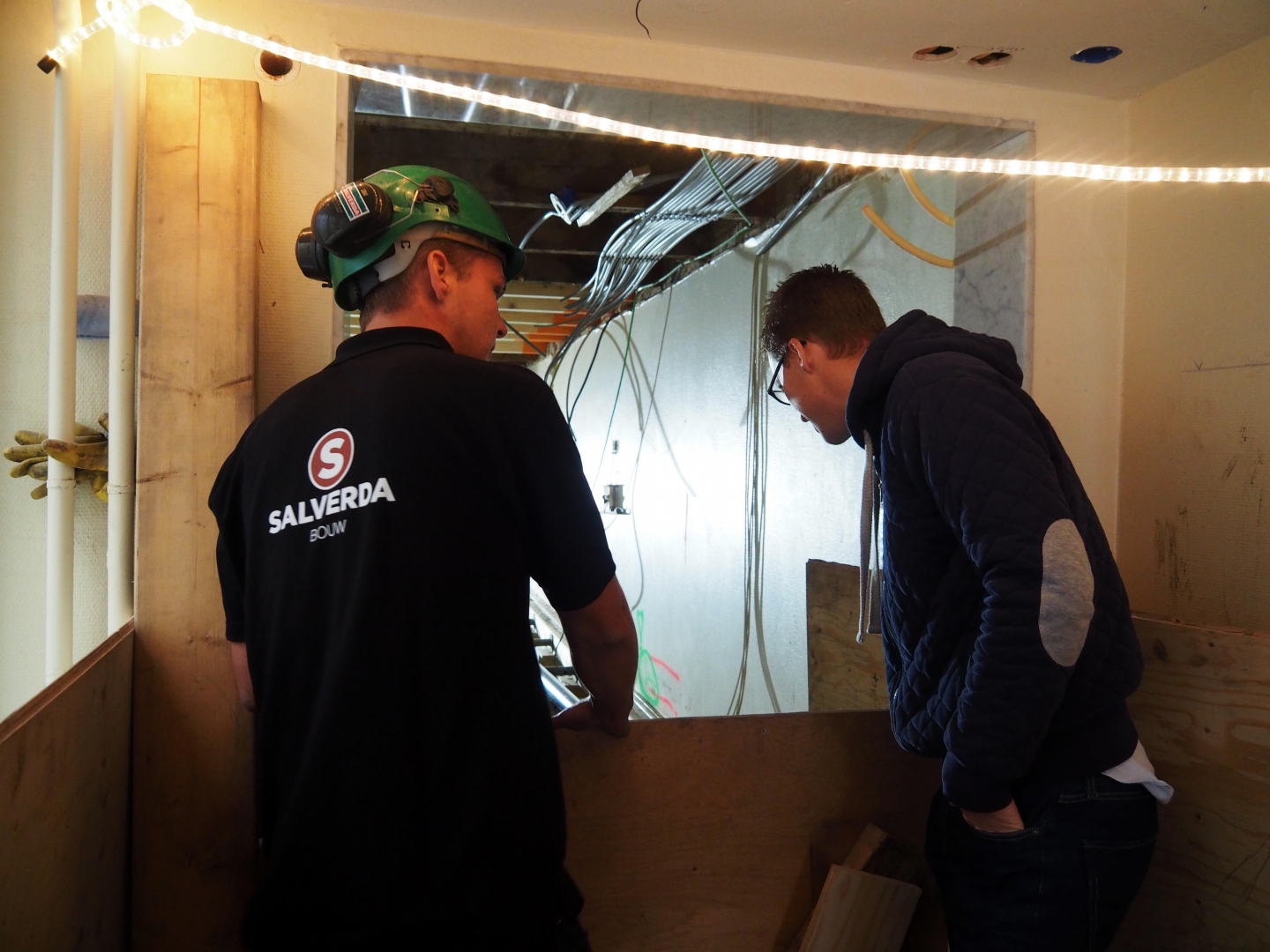 Een medewerker van Salverda Bouw met helm en een collega kijken naar een doorbraak in een binnenmuur waar leidingen doorheen lopen