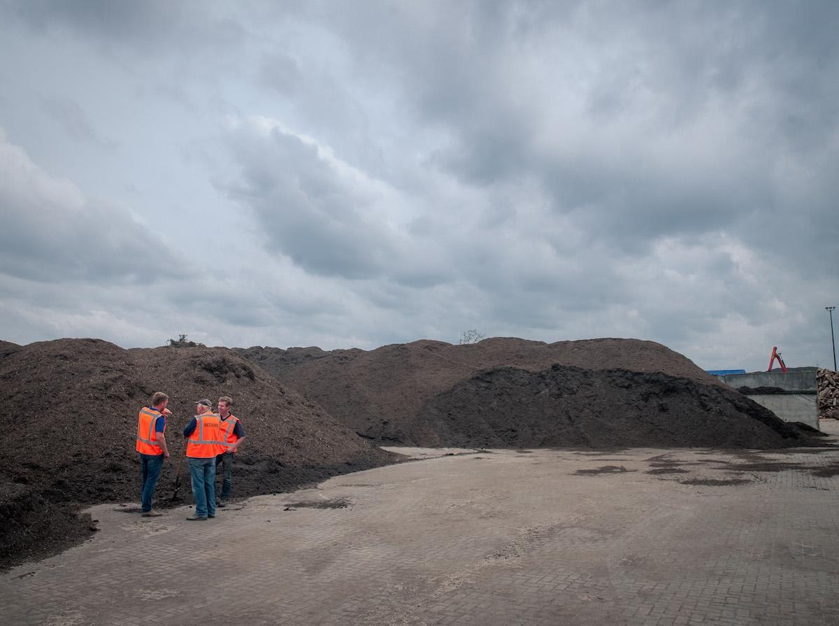 Drie medewerkers in oranje veiligheidsvest tussen lange bergen compost op een recyclinglocatie, onder een bewolkte lucht