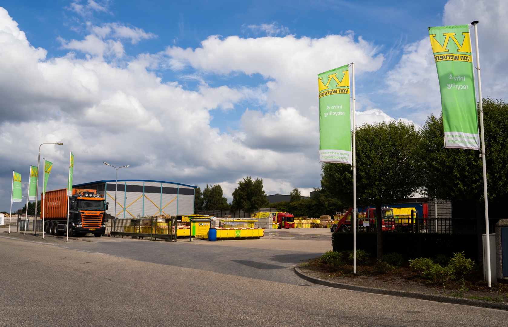 De ingang van de vestiging met groene Van Werven-vlaggen, de blauwe loods en gele containers, met een vertrekkende vrachtwagen