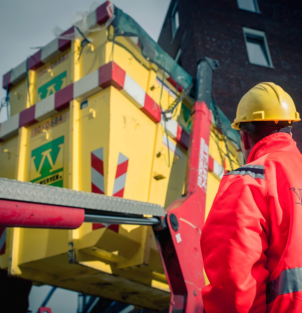 Medewerker van Van Werven kijkt toe terwijl een gele afzetcontainer met een kraan op een vrachtwagen wordt gezet