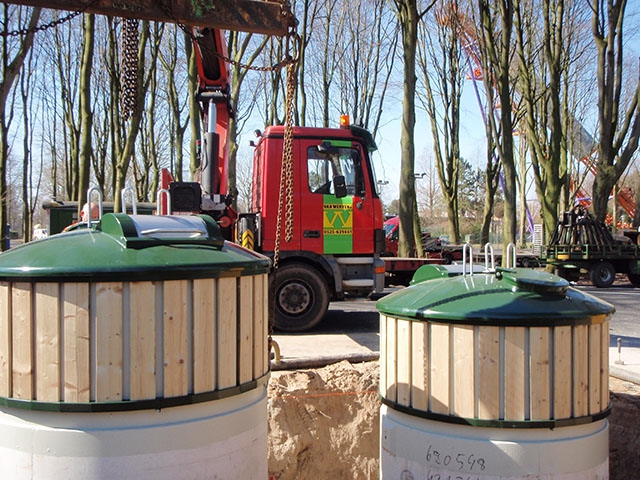Twee semi-ondergrondse containers met houten omkasting worden met een kraanwagen van Van Werven in een uitgegraven put geplaatst