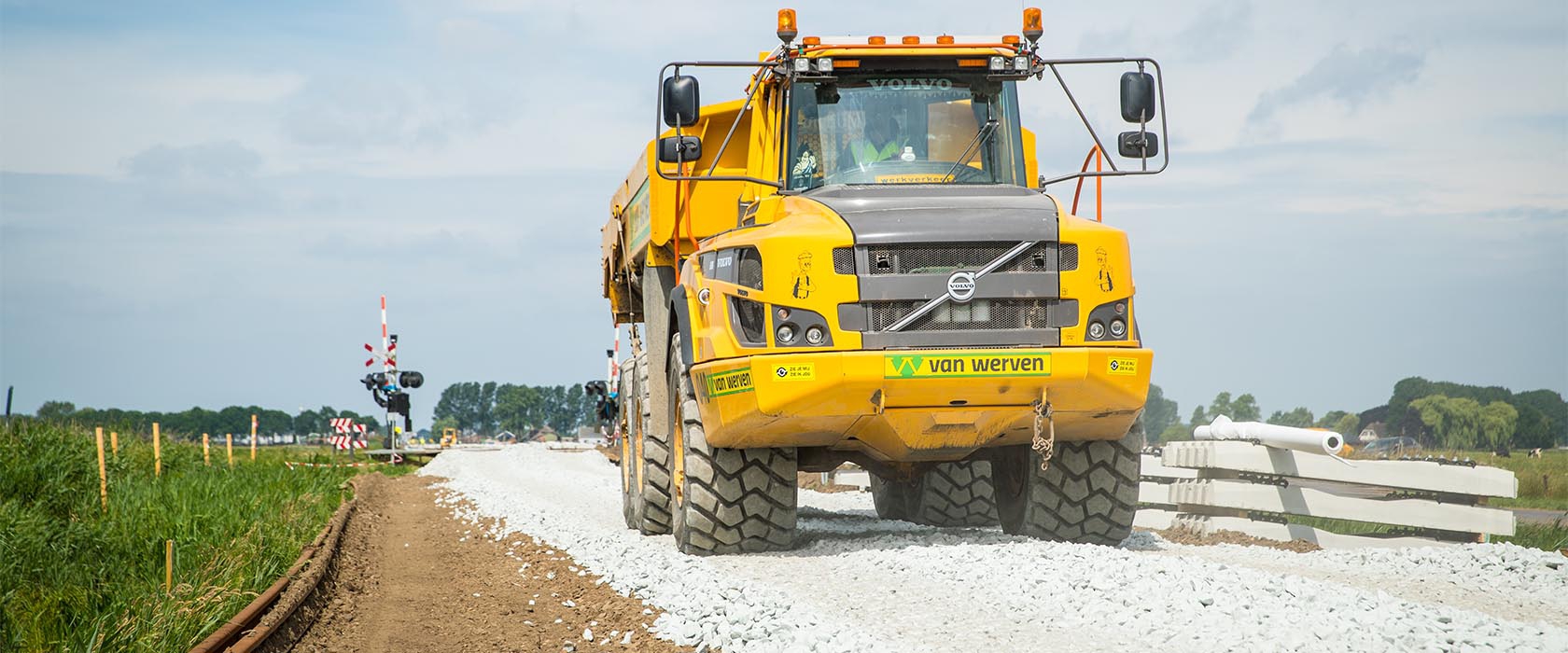 Gele knikdumper van Van Werven rijdt over een pas aangelegde steenslagbaan langs een spoorwegovergang