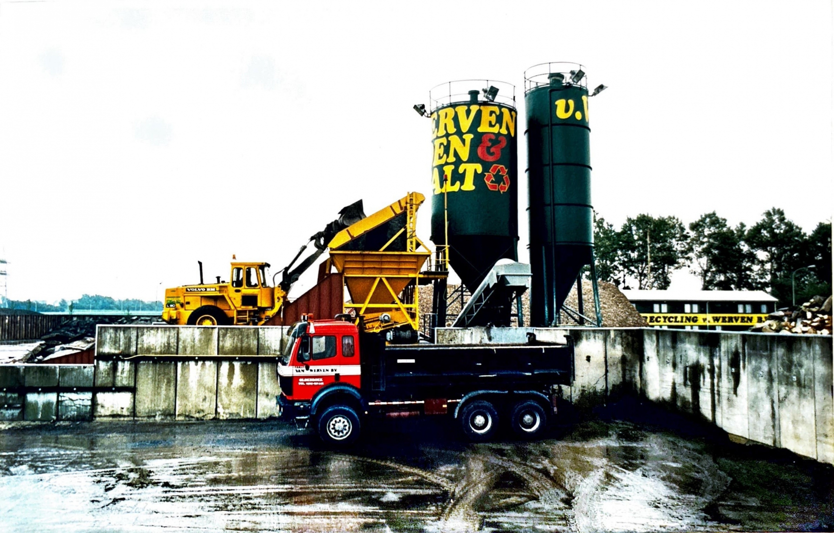 Archieffoto uit de begintijd van de vestiging: een rode kipper van Van Werven en een gele Volvo-shovel bij de groene silo's met het opschrift Van Werven Asfalt en een recyclingsymbool