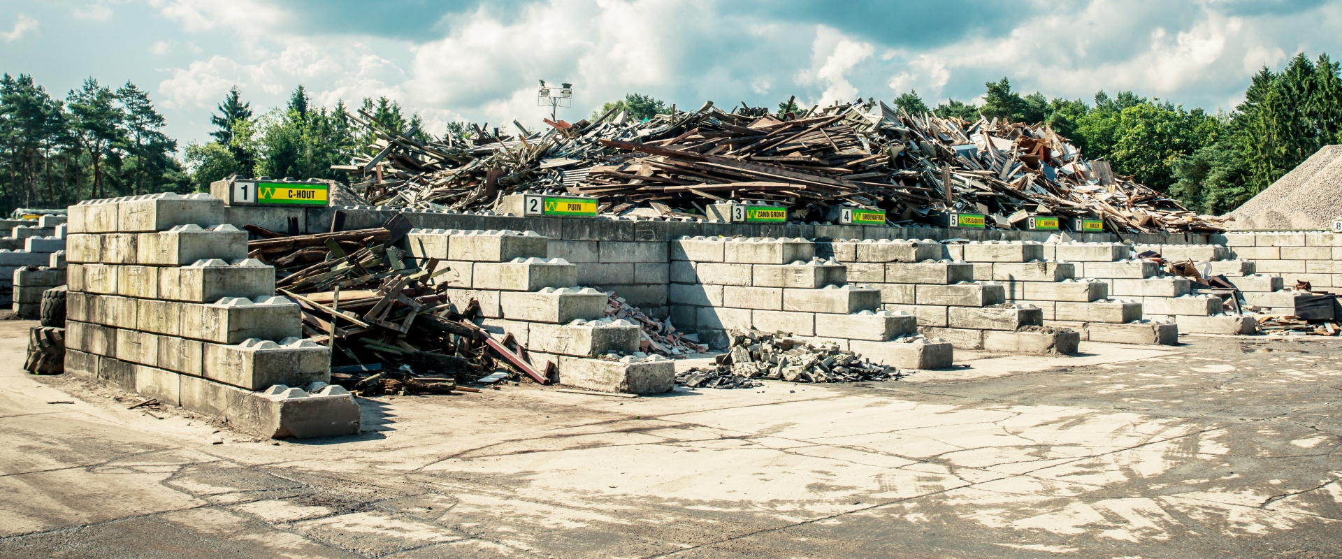 Gescheiden afvalstromen in genummerde vakken van betonblokken op een recyclingvestiging van Van Werven, met daarachter een berg sloophout
