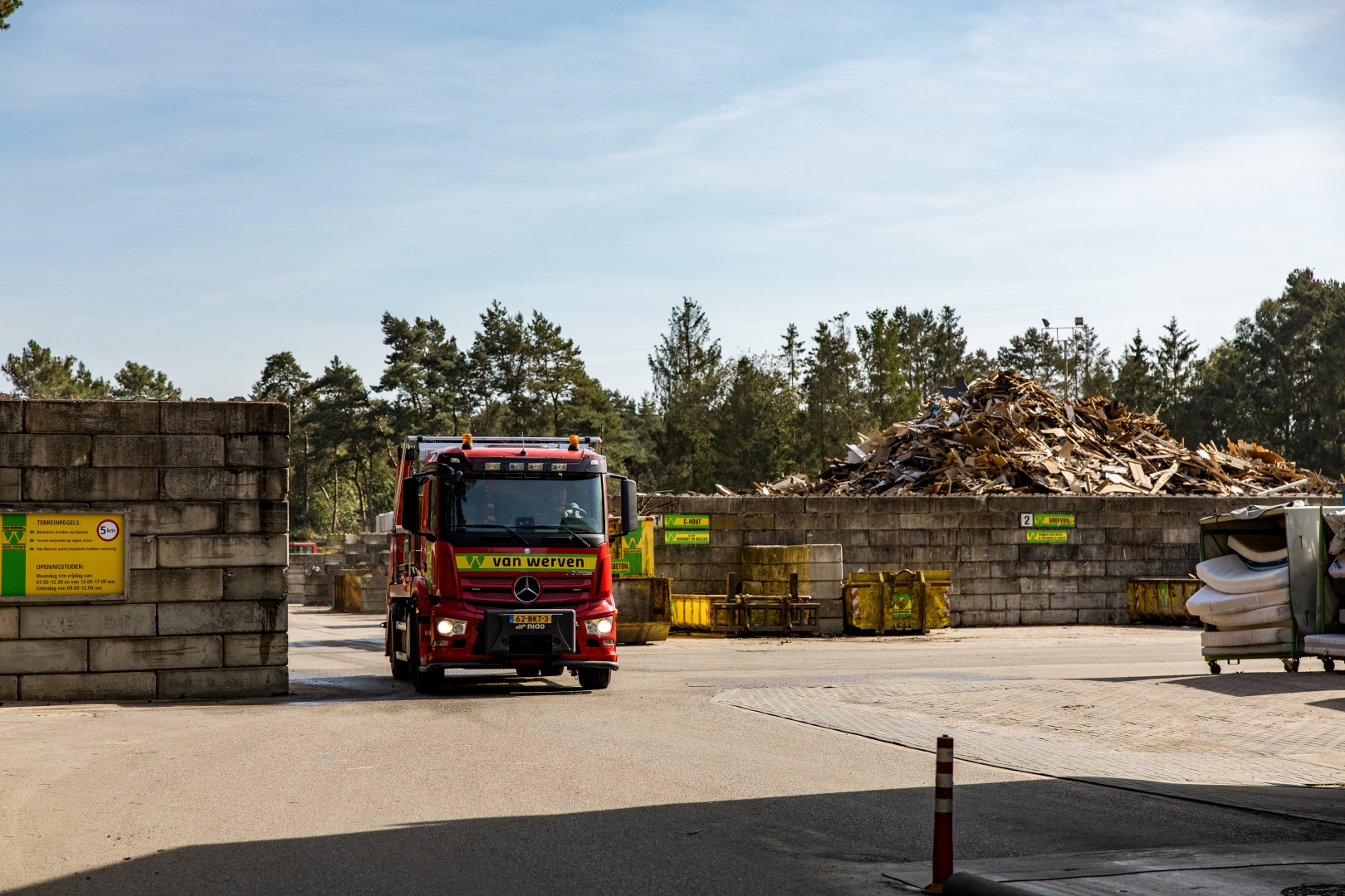 Een rode Van Werven-vrachtwagen rijdt de poort van de recyclinglocatie uit, met een berg houtafval en gele containers op de achtergrond